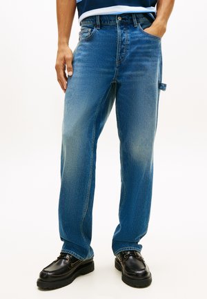 Pieaugušais valkā izbalējušas zilas tiegu bikses un melnas ādas kurpes ar šņorēm, stāvot ar vienu roku kabatā.