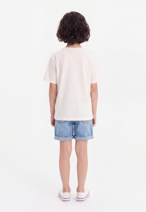 Enfant aux cheveux bouclés debout, de dos, portant un t-shirt blanc uni, un short en jean retroussé et des baskets blanches sur fond blanc.