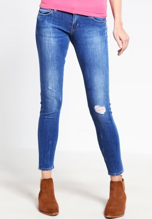 Vrouw die blauwe skinny jeans draagt met een scheur op de rechterknie, bruine suède enkellaarzen en een roze shirt, staand voor een witte achtergrond.