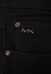 Schwarze Jeans aus Denim besitzen silberne Hardware mit sichtbarem "MK"-Logo und kontrastierenden Nähten. Nahaufnahme der Vorderentasche.
