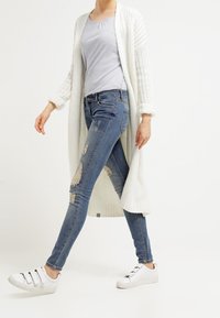 Kevyt valkoinen neulecardigan siniraidallisen paidan päällä, yhdistettynä kulutettuihin sinisiin farkkuihin ja valkoisiin tarranauhatekkereihin.