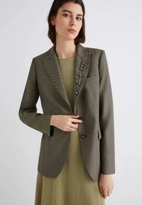 Blazer verde oliva con finitura testurizzata, chiusura a un bottone e decorazioni di perline sulle spalle. Indossato sopra un vestito verde tenue.