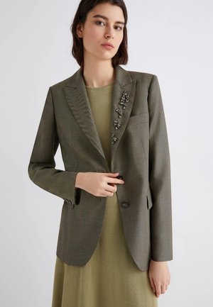Blazer verde oliva con finitura testurizzata, chiusura a un bottone e decorazioni di perline sulle spalle. Indossato sopra un vestito verde tenue.