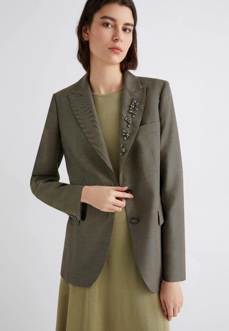 Blazer verde oliva con finitura testurizzata, chiusura a un bottone e decorazioni di perline sulle spalle. Indossato sopra un vestito verde tenue.