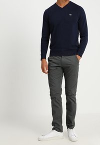 Marineblauer V-Ausschnitt-Pullover mit geripptem Saum und Bündchen, kombiniert mit grauen Slim-Fit-Hosen und weißen Turnschuhen. Auf dem Pullover ist ein gesticktes Logo.
