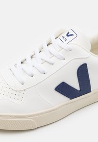 Sneaker bianca in pelle con logo V blu navy, dettagli perforati sulla punta e chiusura con lacci bianchi. Suola in gomma crema con grip testurizzato.