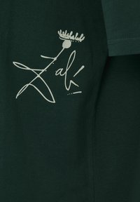 Tissu vert foncé avec un dessin abstrait blanc en lignes représentant une fleur et le texte en cursive "Zak" sur le côté supérieur gauche d'un vêtement.