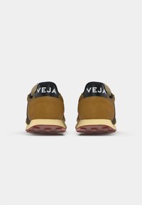Retro in camoscio marrone vista posteriore di scarpe sportive con accenti neri e branding bianco "VEJA"; details della suola intermedia gialla e della suola esterna rossa.