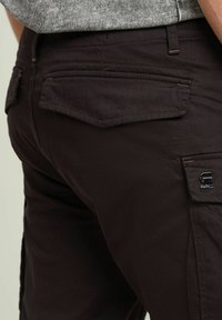 Pantaloni cargo marrone scuro con tasche posteriori piatte, dettagli di cucitura sottili e un piccolo logo sul taschino laterale.