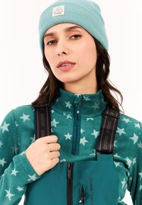 Teal fleece top met sterpatroon, hoge kraag en rits. Zwarte bretels met het label "protest" over een bijpassende teal basislaag.