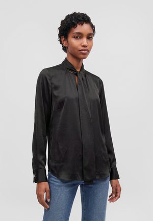 Un mannequin aux cheveux courts et bouclés porte un chemisier noir en satin avec un lien au cou et un jean bleu sur un fond uni.