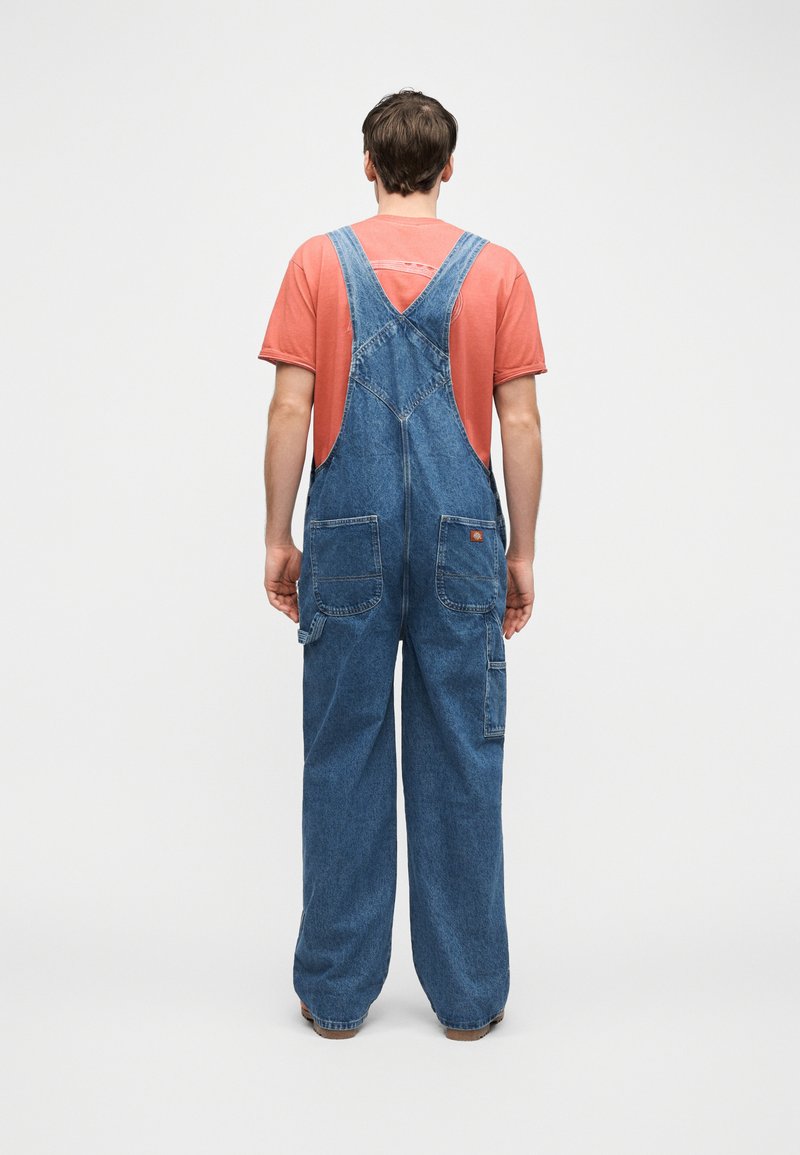 Persona in piedi di spalle che indossa una salopette di denim azzurro chiaro e una maglietta rossa scolorita a maniche corte su uno sfondo uniforme.
