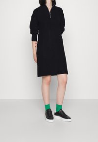 Personne portant une robe en maille noire à manches longues avec une fermeture éclair à l'avant, des chaussettes vertes et des baskets basses noires à semelles blanches.