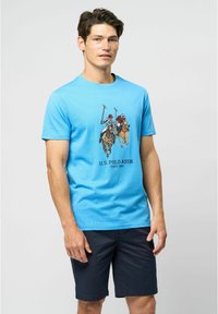Mėlynas medvilninis marškinėlis su užspausdintu grafiką, vaizduojančiu du polo žaidėjus ant žirgų, derinamas su tamsiai mėlynomis šortais, demonstruojamas ant modelio.