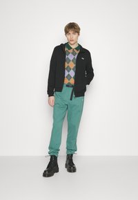 Svart zip-up jakke over en flerfarget argyle-genser, kombinert med grønne bukser og svarte snørebukser. En enkel design med sporty detaljer.