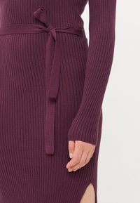 Ribbestrikket kjole i dyp lilla med lange ermer og belte i midjen. Har et sideslitt og glatt tekstur.
