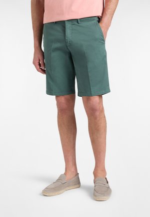 Uomo che indossa pantaloncini verdi fino al ginocchio, camicia rosa e scarpe beige slip-on, in piedi con una mano in tasca su sfondo bianco.