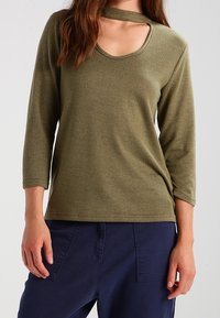 Olijfgroene top met lange mouwen, een ronde halslijn en uitsnijdingen. Gemaakt van zachte stof, met een ontspannen pasvorm en geribde manchetten.
