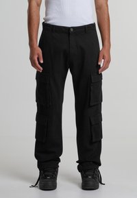 Pantaloni cargo negri, confecționați dintr-un material durabil, cu multiple buzunare laterale, închidere cu nasturi și manșete conice, asortați cu adidași negri.