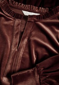 Gros plan d'un chemisier en velours marron avec un col à volants, un décolleté ouvert et des poignets froncés. Une étiquette de vêtement est partiellement visible à l'intérieur du col.