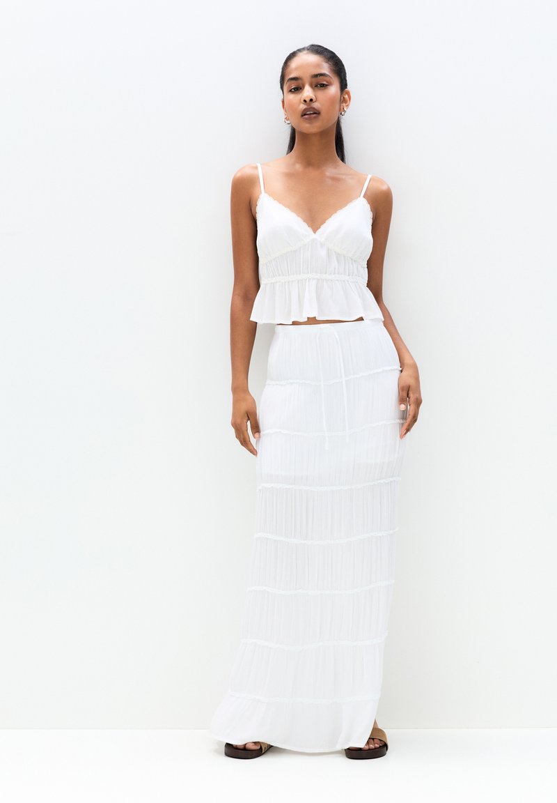 Ensemble blanc en deux pièces comprenant un bralette ajusté avec un ourlet volant et une longue jupe à texture superposée et détails en dentelle.
