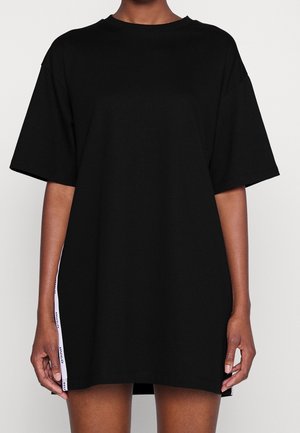 Person iført en løs sort oversized t-shirt med korte ærmer og hvidt brandet bånddetalje på siderne.