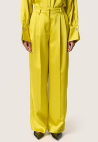 Pantaloni a gamba larga in satin giallo brillante, con vita sartoriale e pieghe strutturate, abbinati a tacchi neri a punta.