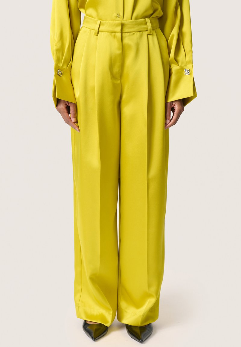 Pantaloni a gamba larga in satin giallo brillante, con vita sartoriale e pieghe strutturate, abbinati a tacchi neri a punta.