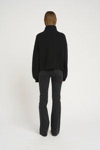 Schwarzer gestrickter Pullover mit hohem Kragen, lässiger Passform und überschnittenen Schultern, kombiniert mit schwarzen Schlagjeans, von hinten betrachtet.