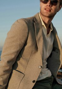 Blazer texturizado gris claro con solapa muesca, cierre de un solo botón y un bolsillo en el pecho sobre una camisa blanca con estampado.