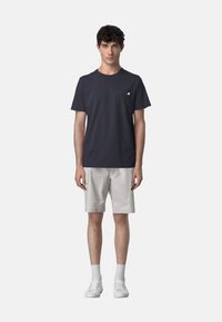 T-shirt in cotone blu navy con una piccola tasca sul petto, abbinata a pantaloni corti grigi chiari. Il modello indossa sneakers bianche e calzini bianchi a coste.