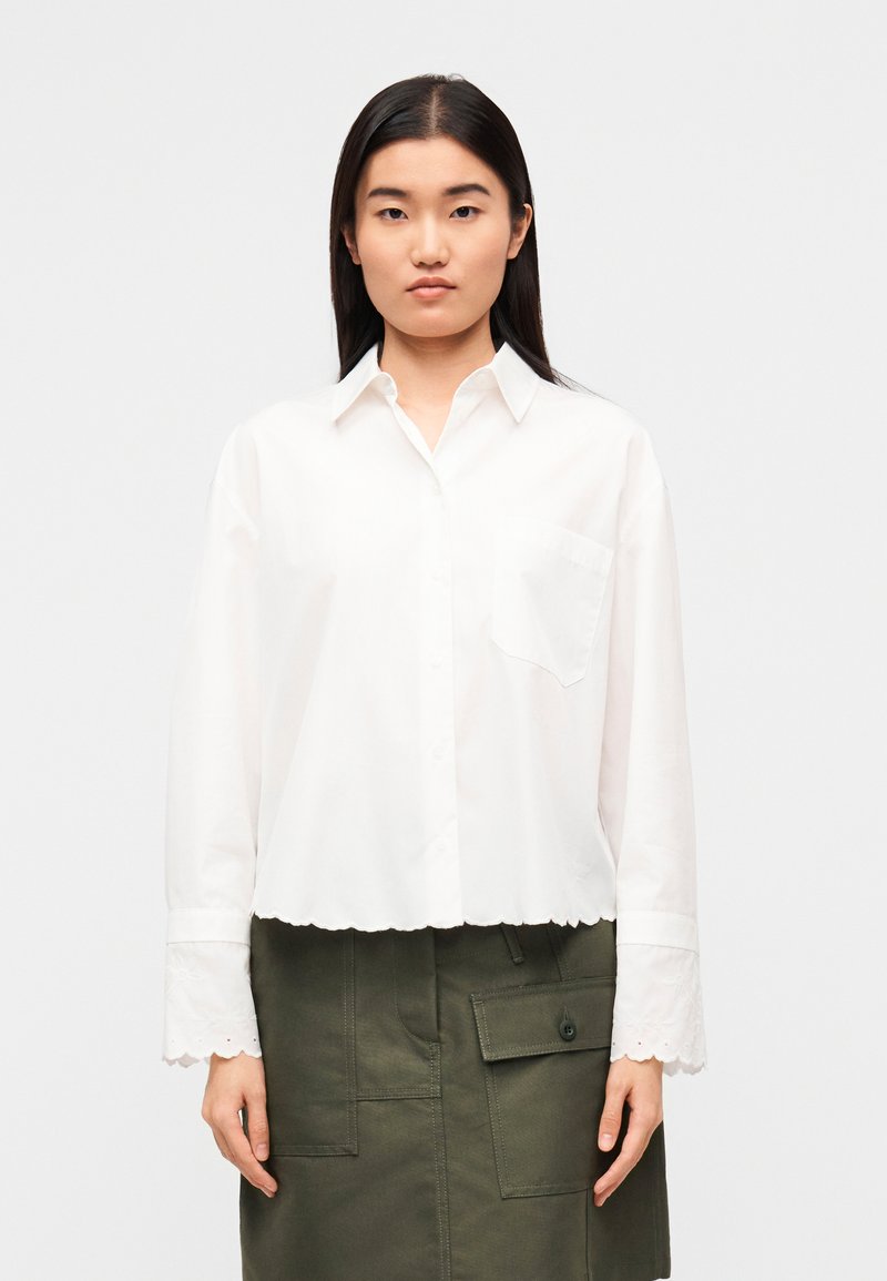 Λευκό κοντό πουκάμισο με κουμπιά και τσέπη, με κυ ματοειδές τελείωμα και μακριά μανίκια με κεντημένες λεπτομέρειες. Συνδυάζεται με πράσινη φούστα.