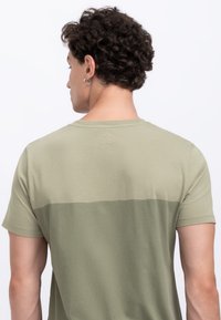 T-shirt verde khaki con maniche corte, caratterizzato da una parte superiore più chiara e dettagli testurizzati nella parte posteriore del collo. Materiale in cotone liscio.
