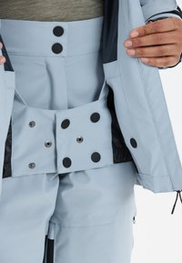 Hellblauer wasserdichter Mantel mit schwarzen Knöpfen und Druckknöpfen, ausgestattet mit einem glatten, strukturierten Stoff und einem verstellbaren Innenfutter.