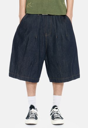 Persona con pantalones cortos holgados de mezclilla oscura hasta la rodilla, combinados con calcetines blancos y zapatillas negras bajas, de pie con las manos en los bolsillos.