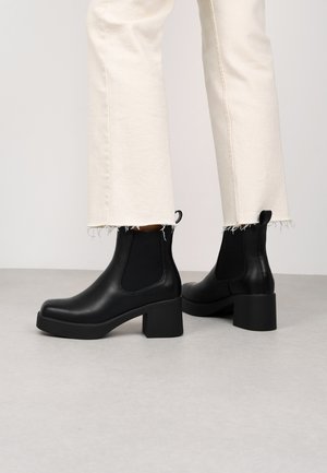 Botas de tobillo de cuero negro con un tacón grueso, paneles laterales elásticos y una tirador en la parte trasera. Combinadas con pantalones blancos con dobladillo deshilachado.