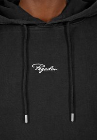 Čierna mikina s kapucňou vyrobená z mäkkej tkaniny; má vyšité biele logo "Pegador" a čierne šnúrky s objednávkami z nehrdzavejúcej ocele.