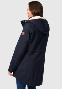 Marineblaues Jackett mit einer gefütterten Kapuze aus Kunstpelz, featuring ein rundes Logo-Patch am Ärmel und einem leicht längeren Rückenbereich.