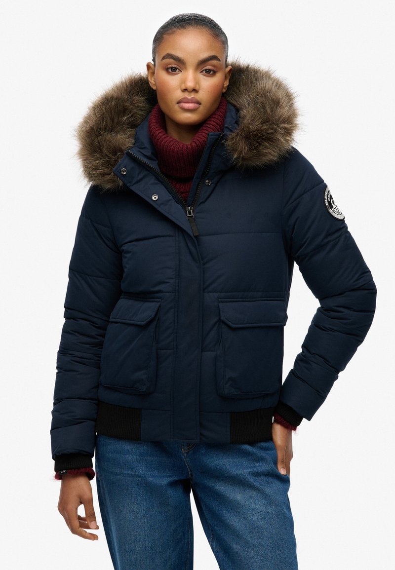 Marineblå puffjakke med en pelsforet hætte, udstyret med to frontlommer og ribbede manchetter, båret over en bordeaux turtleneck sweater.