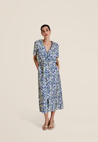 Robe portefeuille à motifs bleus et verts. Manches courtes, décolleté en V profond, taille cintrée avec lien, et poches. Design mi-long.