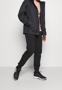 Czarna wodoodporna kurtka, jasnoróżowa koszula, czarne spodnie dresowe oraz czarne buty sportowe z białymi akcentami. Kurtka posiada kaptur i widoczne zamki błyskawiczne.