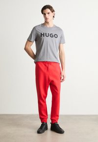 Grijze katoenen T-shirt met grote zwarte "HUGO"-print, gecombineerd met rode trainingsbroeken met elastische tailleband en zwarte sneakers.
