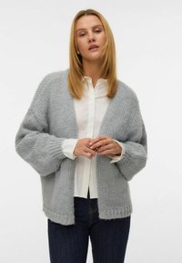 Cárdigan de punto gris claro con un ajuste holgado, mangas anchas y un dobladillo acanalado, llevado sobre una camisa blanca de botones. Hilo texturizado.