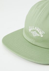Casquette en tissu vert avec une visière plate et un logo brodé blanc. Présente des détails cousus sur la visière et les panneaux latéraux.