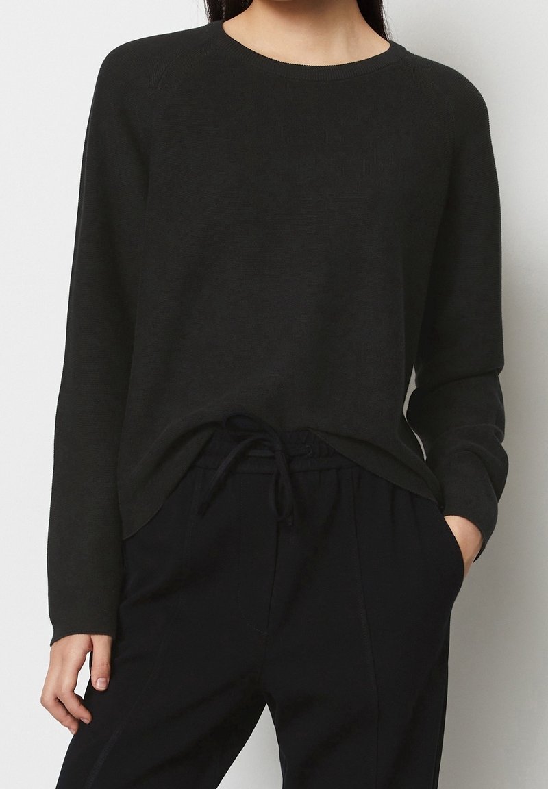 Femme portant un pull noir à manches longues et col rond et un pantalon noir à cordon de serrage, une main dans la poche, sur un fond uni.