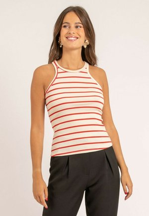 Jeune femme souriante, portant un haut blanc sans manches à rayures horizontales rouges et un pantalon taille haute noir, debout devant un fond uni.