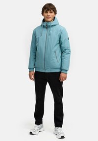 Ljusblå isolerad jacka med huva, framdragkedja och sidofickor. I kombination med svarta byxor och vita sneakers, stående mot en vit bakgrund.