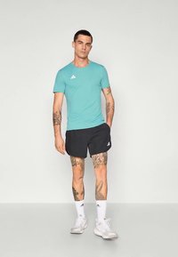 Tyrkysové športové tričko s krátkym rukávom a logom Adidas v kombinácii s čiernymi šortkami, nosené s bielymi ponožkami a topánkami.