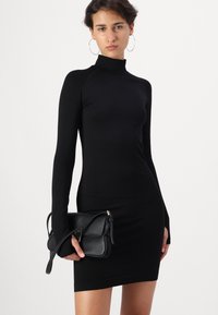 Melna pieguļoša kleita ar garām piedurknēm un augstu apkakli, izgatavota no ribota auduma. Tur rokās nelielu melnu somiņu ar pleca siksnu.