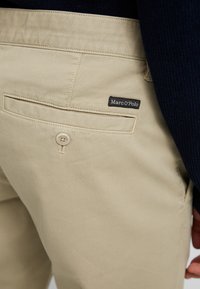Beige katoenen broek met een achterzak met een knop, met een zwart "Marc O'Polo" label. Soepele textuur en rechte pasvorm.